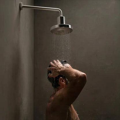 Person washing hair under a shower head in a dimly lit bathroom.